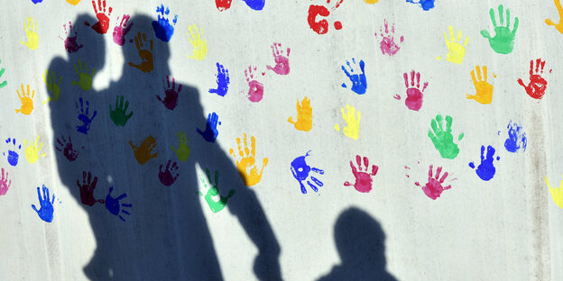 Schatten einer Person, die ein Kind auf dem Arm hat und ein Kind an der Hand hält. Im Hintergrund bunte Handabdrücke von Kindern. Schatten einer Person, die ein Kind auf dem Arm hat und ein Kind an der Hand hält. Im Hintergrund bunte Handabdrücke von Kindern.