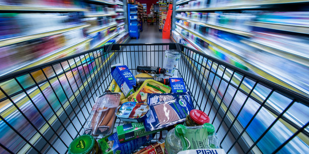 Ein Einkaufswagen mit vielen Lebensmitteln fährt durch einen Supermarkt Ein Einkaufswagen mit vielen Lebensmitteln fährt durch einen Supermarkt