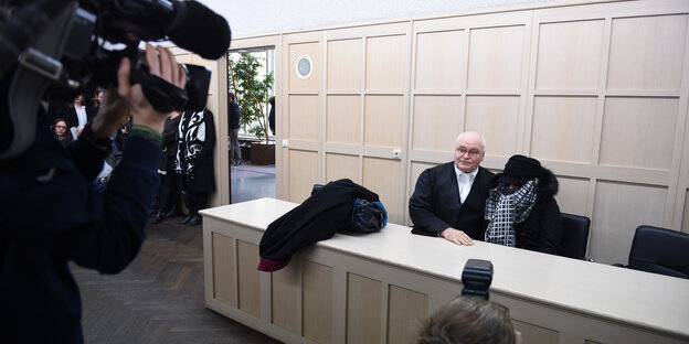 Ein alter Mann mit schwarzem Anzug und weißem Hemd sitzt neben einer verhüllten Frau mit schwarzer Schirmmütze, Sonnenbrille, Anorak und Schal. Vor ihnen ist ein Kameramann, der beide filmt. Ein alter Mann mit schwarzem Anzug und weißem Hemd sitzt neben einer verhüllten Frau mit schwarzer Schirmmütze, Sonnenbrille, Anorak und Schal. Vor ihnen ist ein Kameramann, der beide filmt.