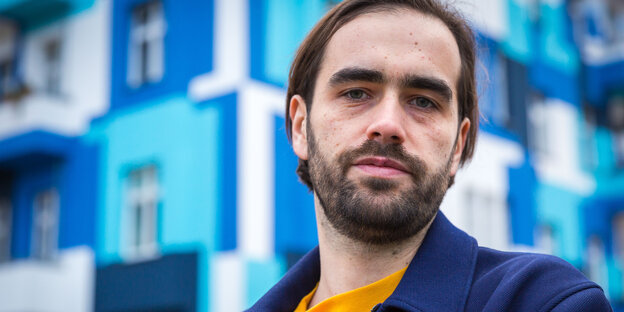 Ein junger Mann im lila Sweatshirt hockt vor einem blauweißen Gebäude Ein junger Mann im lila Sweatshirt hockt vor einem blauweißen Gebäude