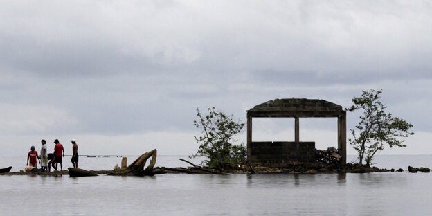 Überschwemmtes Land, fast überall Wasser, einige Menschen stehen neben eingestürztem Haus