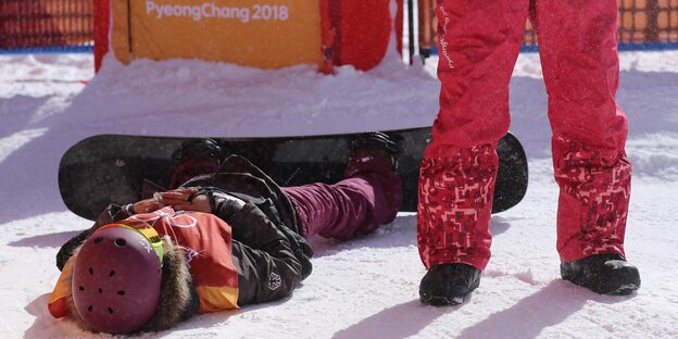 Silvia Mittermüller liegt an ihr Snowboard geschnallt auf dem Schnee. Neben ihr steht jemand in roten Skihosen Silvia Mittermüller liegt an ihr Snowboard geschnallt auf dem Schnee. Neben ihr steht jemand in roten Skihosen