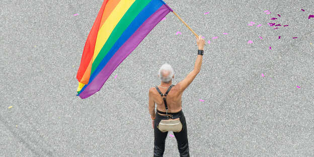 Ein grauhaariger Mann mit freiem Oberkörper und ausgeschnittener Lederhose schwenkt eine Regenbogenfarbe. Ein grauhaariger Mann mit freiem Oberkörper und ausgeschnittener Lederhose schwenkt eine Regenbogenfarbe.