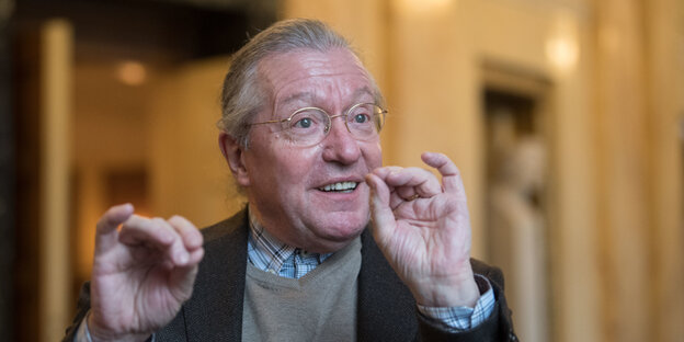 Ein Mann mit grauen Haaren und goldener Brille hebt die Hände und drückt Daumen und Zeigefinger zusammen. Ein Mann mit grauen Haaren und goldener Brille hebt die Hände und drückt Daumen und Zeigefinger zusammen.