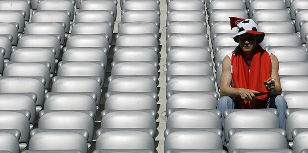Ein Mann mit Fußallhut sitzt ganz allein in den Sitzreihen eines Stadions Ein Mann mit Fußallhut sitzt ganz allein in den Sitzreihen eines Stadions