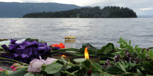 Blumen und Kerzen liegen im Regen am Ufer eines Sees, im Hintergrund ist eine Insel zu sehen