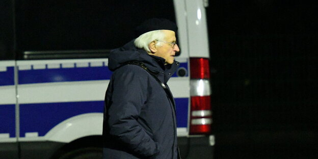 Ein Mann mit grauen Haaren, Baskenmütze, Brille und dunkler Winterjacke geht an einem Kleinbus vorbei.