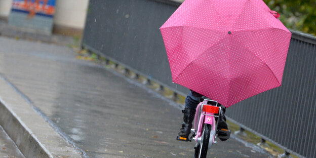 Ein Kind hält einen großen rosafarbenen Regenschirm beim Fahrradfahren über sich (Ansicht von hinten)