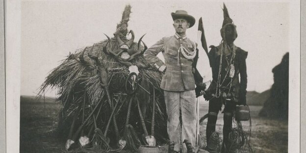 Ein weißter Mann mit dreckiger Uniform steht neben einem schwarzen Mann mit traditioneller Kleidung. Im Hintergrund eine Hütte mit Geweihen. Ein weißter Mann mit dreckiger Uniform steht neben einem schwarzen Mann mit traditioneller Kleidung. Im Hintergrund eine Hütte mit Geweihen.