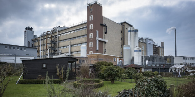 Ein Gartenhaus aus Holz steht auf einem kurz gemähten Rasen. Dahinter ein grau-braunes Fabrikgebäude mit Silos, Rohren und einem rauchenden Schlot. Ein Gartenhaus aus Holz steht auf einem kurz gemähten Rasen. Dahinter ein grau-braunes Fabrikgebäude mit Silos, Rohren und einem rauchenden Schlot.