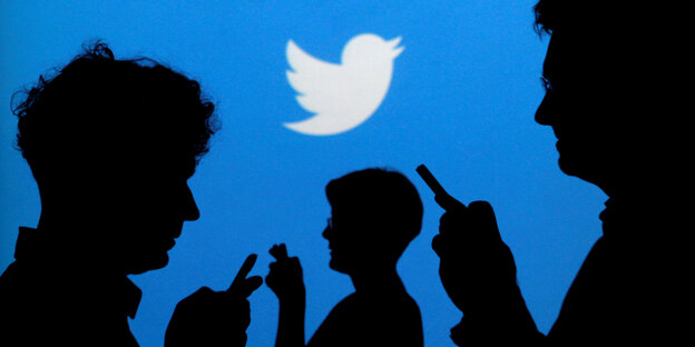 Im Hintergrund eine hellblaue Wand mit einem weißen Vogel-Symbol, im Vordergrund die Umriss von Menschen, die auf ihr Handy tippen Im Hintergrund eine hellblaue Wand mit einem weißen Vogel-Symbol, im Vordergrund die Umriss von Menschen, die auf ihr Handy tippen