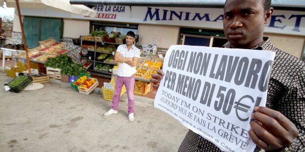 Ein Migrant steht vor einem Laden und demonstriert. Auf seinem Schild steht: "Heute arbeite ich nicht für 50 Euro am Tag". Ein Migrant steht vor einem Laden und demonstriert. Auf seinem Schild steht: "Heute arbeite ich nicht für 50 Euro am Tag".