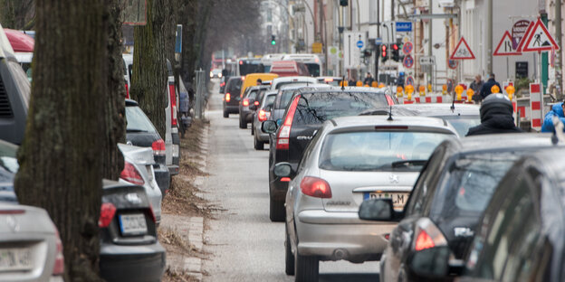 Autos fahren Stoßstange an Stoßstange durch eine Straße. Rechts neben der Autoschlange befindet sich eine Baustelle, links daneben parken Autos.