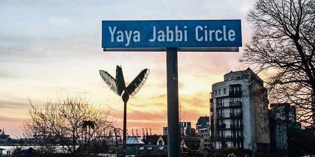 Auf einem Schild steht "Yaya Jabbi Circle" mit weißer Schrift auf blauem Grund Im Hintergrund steht eine künstliche Palme des Park Fiction. Auf einem Schild steht "Yaya Jabbi Circle" mit weißer Schrift auf blauem Grund Im Hintergrund steht eine künstliche Palme des Park Fiction.
