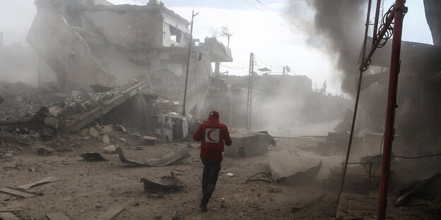 Ein junger Mann von hinten in einem roten Pulli mit einem Halbmond darauf, im Hintergrund Rauchschwaden und zerstörte Gebäude