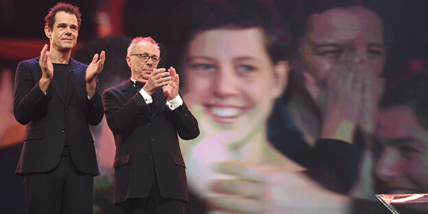 Zwei Männer applaudieren auf einer Bühne, im Hintergrund auf einer Leinwand das überlebensgroße Gesicht einer Frau Zwei Männer applaudieren auf einer Bühne, im Hintergrund auf einer Leinwand das überlebensgroße Gesicht einer Frau