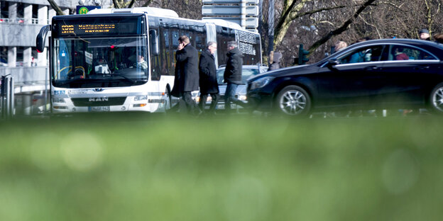 Ein Bus steht an einer Ampel, während Personen vorbei laufen und Autos vorbei fahren.