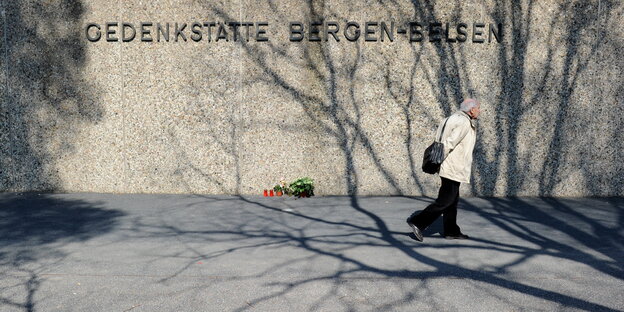 Ein alter Mann geht an einer Mauer vorbei, vor der Blumen liegen. Auf der Mauer steht "Gedenkstätte Bergen-Belsen". Ein alter Mann geht an einer Mauer vorbei, vor der Blumen liegen. Auf der Mauer steht "Gedenkstätte Bergen-Belsen".