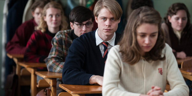 In Schulbänken sitzen Schüler_innen mit ernsten Gesichtern In Schulbänken sitzen Schüler_innen mit ernsten Gesichtern