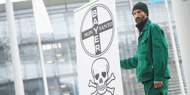 Ein Mann in grüner Kleidung und Mütze hält ein Banner, auf dem die Schriftzüge von Bayer und Monsanto sowie ein Totenschädel zu sehen sind Ein Mann in grüner Kleidung und Mütze hält ein Banner, auf dem die Schriftzüge von Bayer und Monsanto sowie ein Totenschädel zu sehen sind