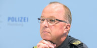 Ein Mann mit hoher Stirn, kurzen grau-blonden Haaren und einer dunklen Brille sitzt vor einer blauen Wand, auf der "Polizei" steht.
