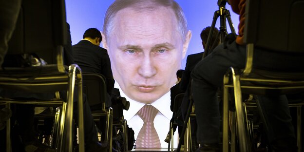 Menschen sitzen in Reihen vor einer Leinwand, auf der Putins Gesicht zu sehen ist Menschen sitzen in Reihen vor einer Leinwand, auf der Putins Gesicht zu sehen ist