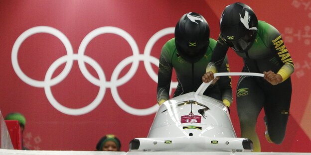 Zwei Bobfaher mit Helmen hintereinander schieben ihren Bob im Laufen an. Im Hintergrund auf einer Wand die olympischen Ringe