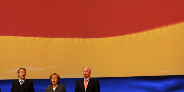 Angela Merkel singt die deutsche Nationalhymne vor einer deutschen Fahne. Neben ihr stehen zwei Parteikollegen Angela Merkel singt die deutsche Nationalhymne vor einer deutschen Fahne. Neben ihr stehen zwei Parteikollegen