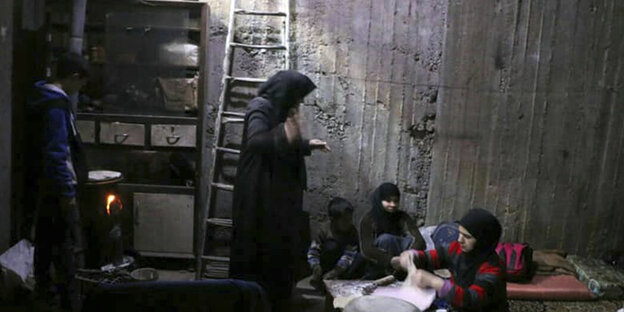 Eine Frau steht in einem Bunker und guckt zu zwei Kinder und einer Frau n, die vor ihr auf dem Boden sitzen Eine Frau steht in einem Bunker und guckt zu zwei Kinder und einer Frau n, die vor ihr auf dem Boden sitzen
