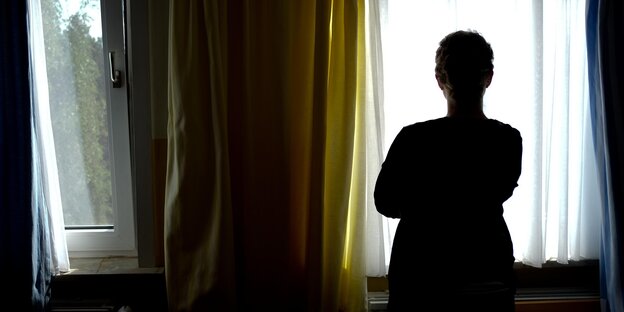 Eine Frau steht am Fenster und schaut hinaus, man sieht nur ihren Rücken Eine Frau steht am Fenster und schaut hinaus, man sieht nur ihren Rücken