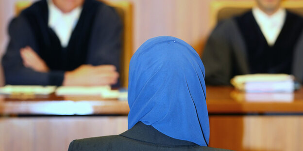 Eine Frau mit blauem Kopftuch ist von hinten zu sehen. Vor ihr sieht man unscharf einen Richter und eine weitere Person Eine Frau mit blauem Kopftuch ist von hinten zu sehen. Vor ihr sieht man unscharf einen Richter und eine weitere Person