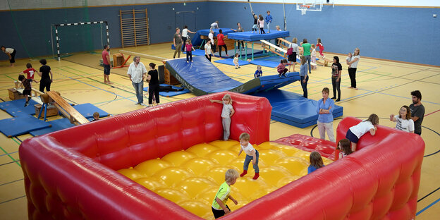 Viele Kinder toben in einer Turnhalle herum und springen auf einer Hüpfburg und stehen auf einer Wippe Viele Kinder toben in einer Turnhalle herum und springen auf einer Hüpfburg und stehen auf einer Wippe