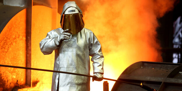 Ein Stahlarbeiter in Schutzkleidung mit Helm steht vor eine lodernden Feuer Ein Stahlarbeiter in Schutzkleidung mit Helm steht vor eine lodernden Feuer