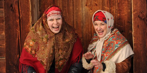 Zwei Frauen in traditionell russicher Kleidung sitzen vor einer Holzwand und lachen Zwei Frauen in traditionell russicher Kleidung sitzen vor einer Holzwand und lachen