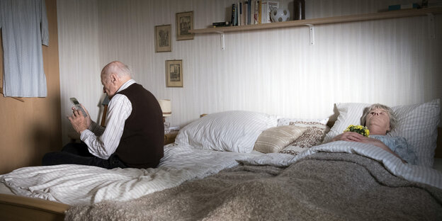 Ein alter Mann sitzt auf einem Ehebett und hält ein Telefon in der Hand. Auf der anderen Seite des Bettes liegt eine Frau mit geschlossenen Augen, die ein Blumensträuschen in den Händen hält Ein alter Mann sitzt auf einem Ehebett und hält ein Telefon in der Hand. Auf der anderen Seite des Bettes liegt eine Frau mit geschlossenen Augen, die ein Blumensträuschen in den Händen hält