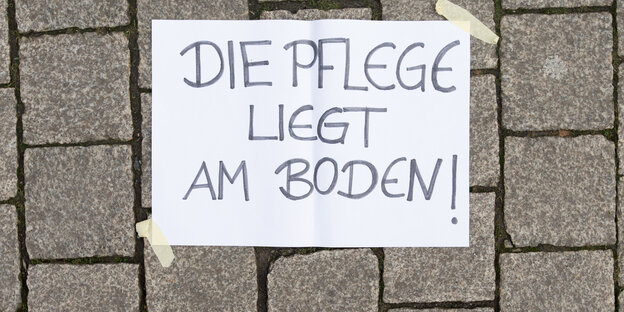 Ein Papier klebt auf Plastersteinen. Aufschrift: Die Pflege liegt am Boden! Ein Papier klebt auf Plastersteinen. Aufschrift: Die Pflege liegt am Boden!