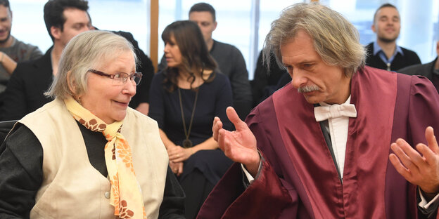 Eine ältere Dame mit Halstuch spricht mit einem Mann neben ihr, der eine rote Anwaltsrobe trägt Eine ältere Dame mit Halstuch spricht mit einem Mann neben ihr, der eine rote Anwaltsrobe trägt