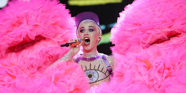 Katy Perry singt in ein Mikrofon, neben ihr rosafarbene Stoffberge Katy Perry singt in ein Mikrofon, neben ihr rosafarbene Stoffberge