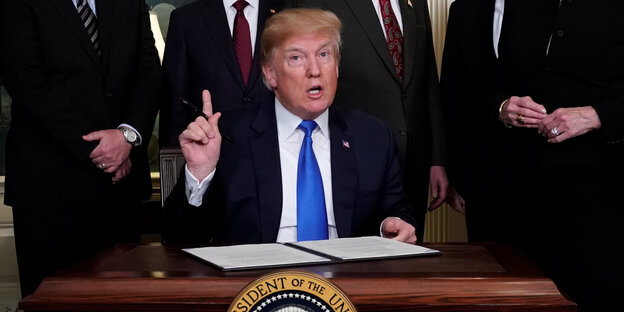 Trump sitzt an einem Schreibtisch und hält den Zeigefinger hoch Trump sitzt an einem Schreibtisch und hält den Zeigefinger hoch