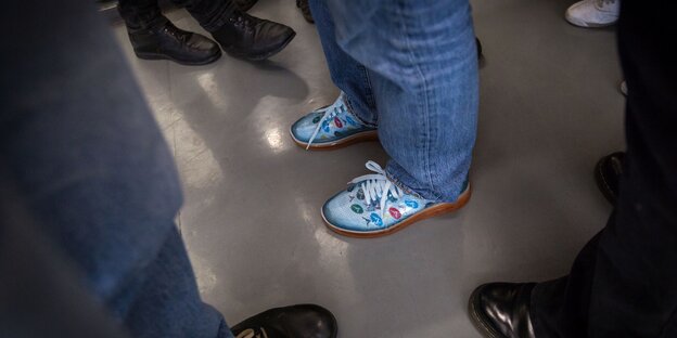 Alle Fußpaare tragen schwarze Schuhe. In der Mitte die Füße von Deniz mit seinen blau-gemusterten Schuhen. Alle Fußpaare tragen schwarze Schuhe. In der Mitte die Füße von Deniz mit seinen blau-gemusterten Schuhen.