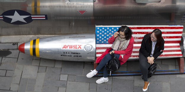 zwei Frauen sitzen auf einer mit einer US-Flagge dekorierten Bank in Form eines Sprengkopfes zwei Frauen sitzen auf einer mit einer US-Flagge dekorierten Bank in Form eines Sprengkopfes