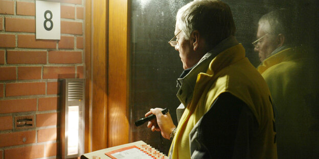 Ein Postbote mit einem Päckchen in der Hand leuchtet mit einer Taschenlampe auf eine Hausklingel Ein Postbote mit einem Päckchen in der Hand leuchtet mit einer Taschenlampe auf eine Hausklingel