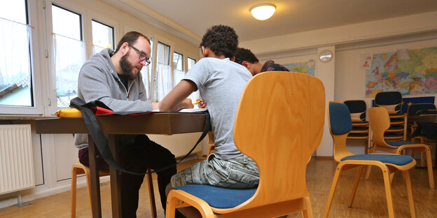 Ein junger Mann mit Bart und Brille arbeitet mit zwei Jugendlichen an einem Tisch. Alle lösen eine Aufgabe. Ein junger Mann mit Bart und Brille arbeitet mit zwei Jugendlichen an einem Tisch. Alle lösen eine Aufgabe.