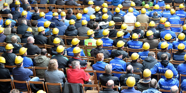 Etliche Mitarbeiter der Meyer-Werft sitzen in Arbeitskleidung auf Stühlen und blicken auf ein Podium. Etliche Mitarbeiter der Meyer-Werft sitzen in Arbeitskleidung auf Stühlen und blicken auf ein Podium.