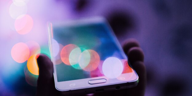 Eine Hand hält ein Smartphone, bunte Lichtreflexionen scheinen auf Eine Hand hält ein Smartphone, bunte Lichtreflexionen scheinen auf