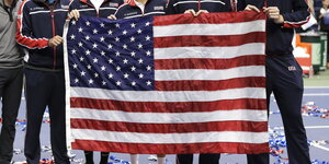 fünf Sportler in Trainingsanzügen halten in einem Stadion eine große US-Fahne in den Händen