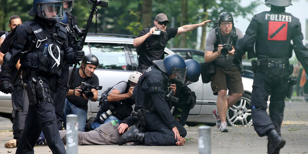 Zwei Polizisten mit Helmen hocken auf einem Demonstranten und drücken ihn mit den Knien zu Boden. Zwei Polizisten mit Helmen hocken auf einem Demonstranten und drücken ihn mit den Knien zu Boden.