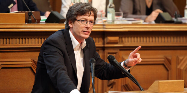 Ein mittelalter Mann steht an einem Rednerpult und hebt den linken Zeigefinger. Ein mittelalter Mann steht an einem Rednerpult und hebt den linken Zeigefinger.