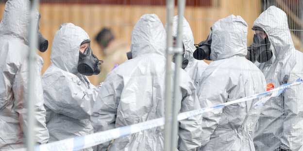 Eine Gruppe Menschen in weißen Schutzanzügen hinter einem Absperrband Eine Gruppe Menschen in weißen Schutzanzügen hinter einem Absperrband