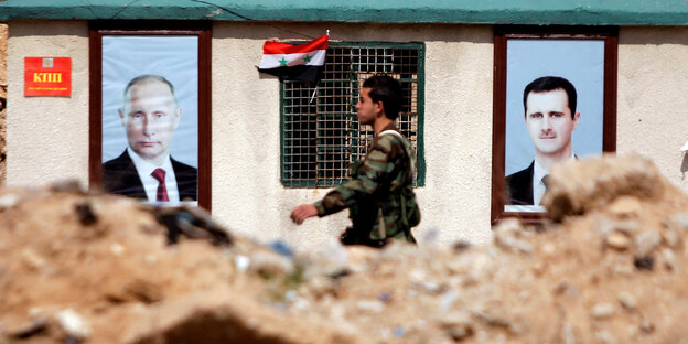 Ein Soldat läuft an einem Haus entlang. Daran kleben Poster mit den Portraits von Putin und Assad Ein Soldat läuft an einem Haus entlang. Daran kleben Poster mit den Portraits von Putin und Assad
